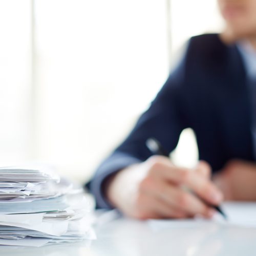 Stack of documents at workplace and male employee on background